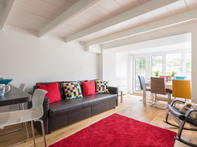 A living room with a black leather sofa with colorful cushions a red rug and a dining area with multicolored chairs at Beehive Cottage in Port Isaac