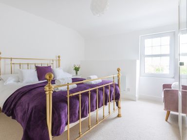 A bedroom with a gold metal bed frame purple bedding white pillows a window and a pink chair at Beehive Cottage in Port Isaac