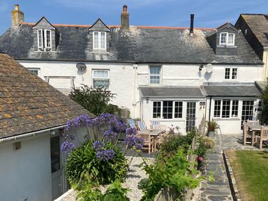 The back view of a white cottage with a slate roof a garden with flowers wooden outdoor tables and chairs and a stone path at Beehive Cottage in Port Isaac