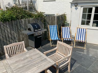 An outdoor patio with a wooden table and chairs a gas barbecue and three striped deck chairs at Beehive Cottage in Port Isaac