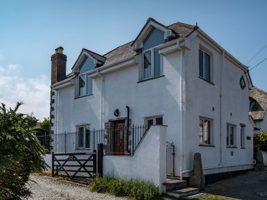 A white two-story house with a chimney and multiple windows surrounded by a black metal fence and gate at Fairholme in Port Isaac