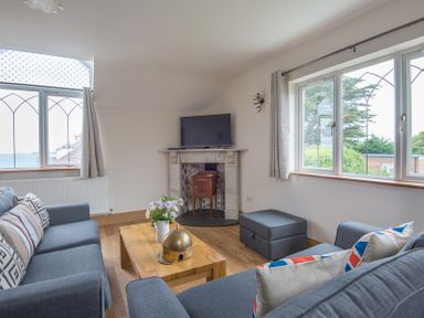A living room with two blue sofas a wooden coffee table flowers on the table a TV on a fireplace and windows with curtains at Fairholme in Port Isaac