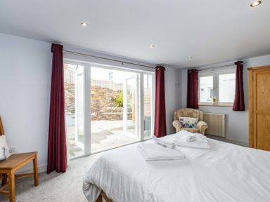 A bedroom with a bed towels a wooden chair an armchair with a cushion a wooden wardrobe and glass doors leading outside at The Hideaway in Port Isaac