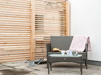 A gray wicker patio loveseat with a pink blanket on the arm and a glass top table with mugs and a teapot on a tiled patio with a wooden privacy screen
