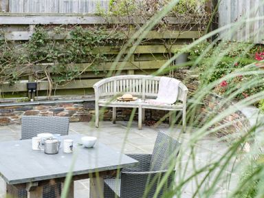 An outdoor patio with a stone floor wicker chairs a table with cups and a teapot a wooden bench with a blanket and hat against a wooden fence at The Hideaway in Port Isaac