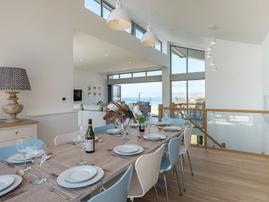 A dining table set with plates glasses and cutlery in a modern room with windows showing a sea view at Point Break in Polzeath