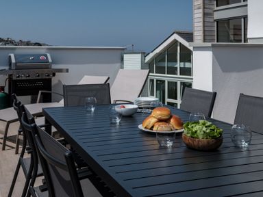 An outdoor dining table with bread rolls lettuce and empty glasses on a terrace with chairs and a barbecue at Point Break in Polzeath