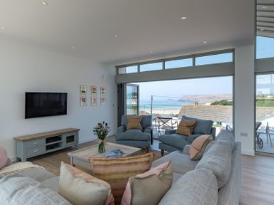 A living room with sofas and armchairs facing a coffee table with flowers and a TV on a stand with a view of the sea at Point Break in Polzeath