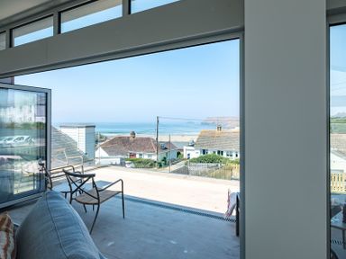 A seating area with two chairs and a sofa facing a large open window with a view of rooftops and the sea at Point Break in Polzeath