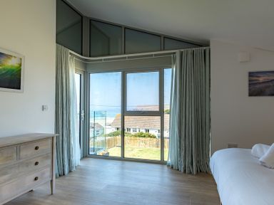 A bedroom with a dresser and a bed near large windows with curtains and a view of houses and the sea at Point Break in Polzeath