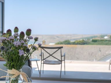 A vase with flowers on a table near a window with a view of a chair and landscape at Point Break in Polzeath