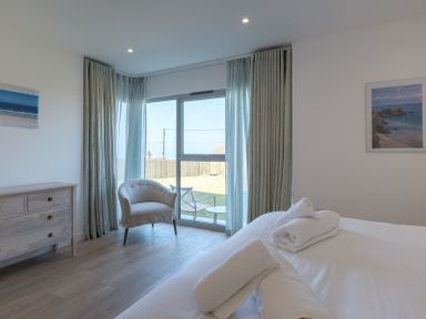 A bedroom with a bed with towels on it an armchair a dresser and large windows with curtains at Point Break in Polzeath