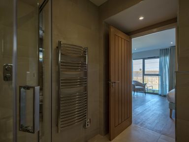 A bathroom with a glass shower enclosure and a heated towel rail leading to a bedroom with a chair and large window at Point Break Polzeath