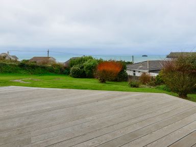 A garden with bushes and grass and a wooden deck overlooking houses and the sea at Sea Mist in Polzeath