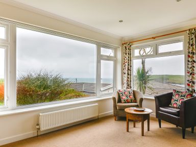 A corner seating area with two armchairs and a wooden round table near large windows with a view of trees and the sea at Sea Mist in Polzeath