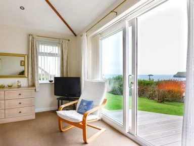 A room with a white cushioned chair and a television near a sliding glass door looking out to a garden and sea view at Sea Mist in Polzeath