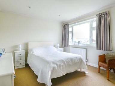 A bedroom with a bed with white bedding two bedside tables and lamps a wicker chair and a window with curtains at Sea Mist in Polzeath