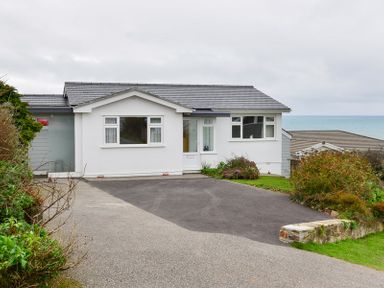 A white single-story house with a driveway and garden with bushes near the sea at Sea Mist in Polzeath
