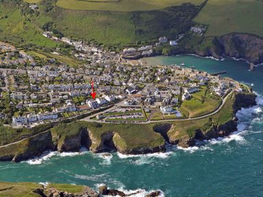 An aerial view of a coastal village with houses, roads, parking lots, green fields, cliffs, and ocean water at The Crows Nest in Port Isaac