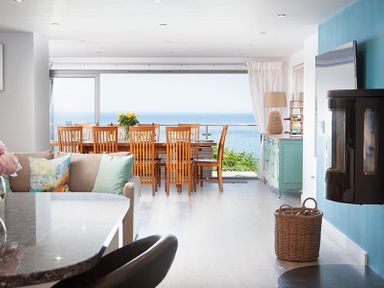 An open living and dining area with wooden chairs at a dining table by large windows showing an ocean view