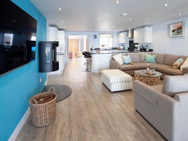 An open plan kitchen and living room with a large sectional sofa a striped ottoman a round coffee table a wall mounted TV and kitchen island with bar stools at The Crows Nest in Port Isaac