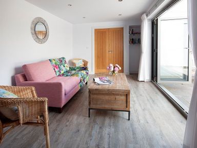A living room with a pink and floral sofa a wicker chair a wooden coffee table with books and flowers and folded wooden doors at The Crows Nest in Port Isaac