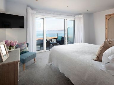 A bedroom with a bed a blue chair a dresser and a balcony overlooking the sea at The Crows Nest in Port Isaac