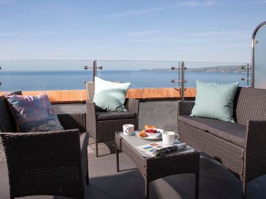 An outdoor seating area with a sofa chairs a table cups and a bowl of fruit overlooking the sea at The Crows Nest in Port Isaac