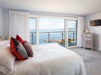 A bedroom with a bed and pillows facing a balcony with ocean view and a small dresser with a television on the wall at The Crows Nest in Port Isaac