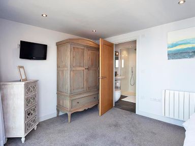 A bedroom with a wooden wardrobe a chest of drawers a wall-mounted TV and a door leading to a bathroom at The Crows Nest in Port Isaac