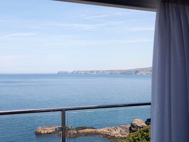 A balcony with a metal railing overlooking the ocean with rocky coastline and a partly curtained window at The Crows Nest in Port Isaac