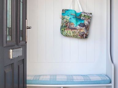 An entryway with a black door a white paneled wall with a hanging colorful tote bag and a blue plaid cushioned bench at The Crows Nest in Port Isaac
