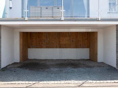An empty gravel garage with wooden back wall under a balcony with glass railing at The Crows Nest in Port Isaac