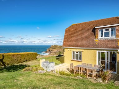 A yellow house with a brown roof next to a patio with wooden chairs and a table facing the sea at Trevan House Lundy in Polzeath