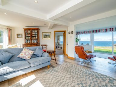 A living room with blue sofa wooden bookcase chairs and large windows showing ocean view at Trevan House Lundy in Polzeath