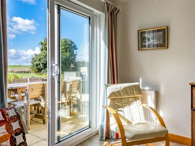 A room with a cushioned chair near glass sliding doors overlooking an outdoor wooden table and chairs at Trevan House Lundy in Polzeath
