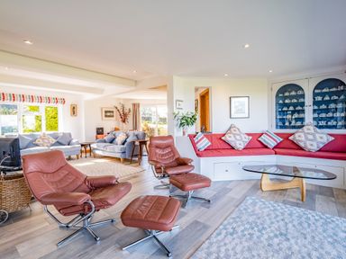 A living room with leather chairs, grey sofas, a built-in red bench with cushions, glass coffee table, and display cabinet at Trevan House Lundy in Polzeath