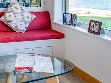 A corner seating area with a red cushion and a patterned pillow next to a window with framed photographs and a glass table with a map on top at Trevan House Lundy in Polzeath