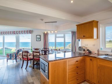 A kitchen and dining area with wooden cabinets a granite countertop dining table with chairs and large windows overlooking the sea at Trevan House Lundy in Polzeath