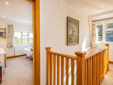 A hallway with wooden railing and a painting on the wall next to a bedroom with a bed and windows with floral curtains at Trevan House Lundy in Polzeath