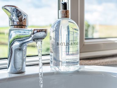 A bathroom sink with running water and a bottle of Baylis and Harding hand wash by a window at Trevan House Lundy in Polzeath