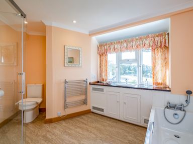 A bathroom with a toilet, bathtub, sink under a window with curtains, a heated towel rail, and a wall mirror at Trevan House Lundy in Polzeath