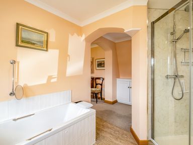 A bathroom with a bathtub and a glass shower enclosure connected to a room with a chair and cabinet at Trevan House Lundy in Polzeath