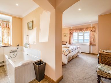 A bedroom with a large bed and a window with curtains next to a bathroom with a bathtub and a wicker bin at Trevan House Lundy in Polzeath