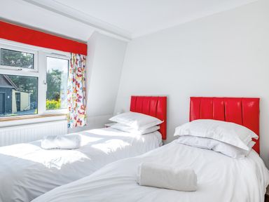A bedroom with two single beds with red headboards and white bedding next to a window with colorful curtains at Trevan House Lundy in Polzeath