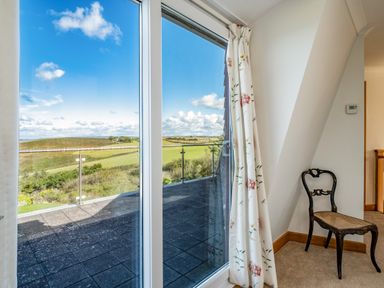 A sliding glass door with floral curtains leading to a balcony with a view of fields and hills and a wooden chair in a carpeted room at Trevan House Lundy in Polzeath