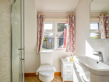 A bathroom with a toilet, a window with patterned curtains, a small drawer unit, and a sink with a mirror at Trevan House Lundy in Polzeath