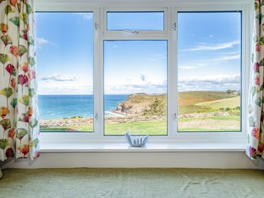 A window with floral curtains overlooking a coastal landscape with cliffs and ocean at Trevan House Lundy in Polzeath