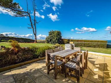 An outdoor patio with a wooden table and chairs near a grassy area with trees and bushes at Trevan House Lundy in Polzeath