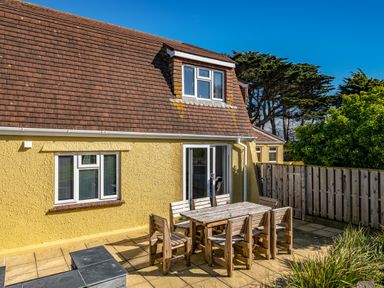An outdoor patio area with wooden table and chairs next to a yellow house with a tiled roof and sliding glass doors at Trevan House Lundy in Polzeath